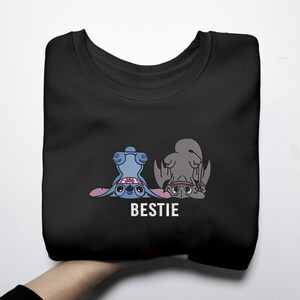 K&ouml;nnte beinhalten: Schwarzes Sweatshirt mit einer Cartoon-Grafik von zwei Figuren, eine blaue und eine graue, mit dem Wort "BESTIE" in Wei&szlig;. Die Figuren liegen und schauen den Betrachter an.