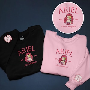 May include: Black and pink sweatshirts with the word "ARIEL" in pink script, a mermaid graphic, and the text "EST. 1989 LITTLE MERMAID." A circular design with the same graphic is visible. The pink sweatshirt sleeve has a "LINDA" patch.