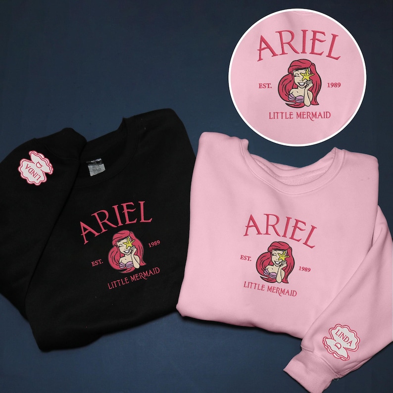 May include: Black and pink sweatshirts with the word "ARIEL" in pink script, a mermaid graphic, and the text "EST. 1989 LITTLE MERMAID." A circular design with the same graphic is visible. The pink sweatshirt sleeve has a "LINDA" patch.