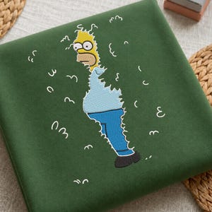 Op de afbeelding: Een donkergroene sweater met een geborduurde afbeelding van een geel stripfiguur dat uit een heg tevoorschijn komt. Het figuur draagt een lichtblauw shirt, een blauwe broek en zwarte schoenen, omringd door witte gestikte contouren van bladeren.