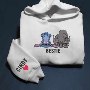 K&ouml;nnte beinhalten: Wei&szlig;er Kapuzenpullover mit langen &Auml;rmeln. Auf der Vorderseite sind eine blaue Zeichentrickfigur und eine graue Zeichentrickfigur abgebildet, darunter das Wort "BESTIE". Der linke &Auml;rmel tr&auml;gt den Namen "CINDY" und ein rotes Herz.
