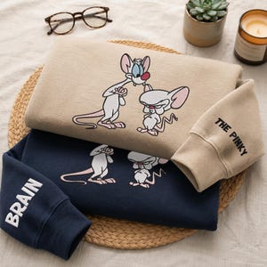 Puede incluir: Una sudadera beige con un gráfico bordado de Pinky y Cerebro, con el texto THE PINKY en la manga, sobre una sudadera azul marino con el mismo gráfico y el texto BRAIN bordado en el puño de la manga.
