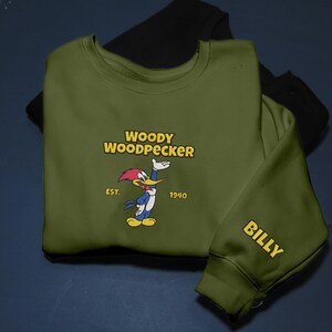 May include: Olive green sweatshirt featuring the Woody Woodpecker cartoon character. The design includes the text "WOODY WOODPECKER" above the cartoon image, with "EST. 1940" below. The sleeve has the name "BILLY" in yellow.