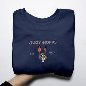 May include: A navy blue sweatshirt with an embroidered design of Judy Hopps, a cartoon rabbit. The text "JUDY HOPPS", "EST", and "2016" are also embroidered. The design is centered on the front of the folded sweatshirt.