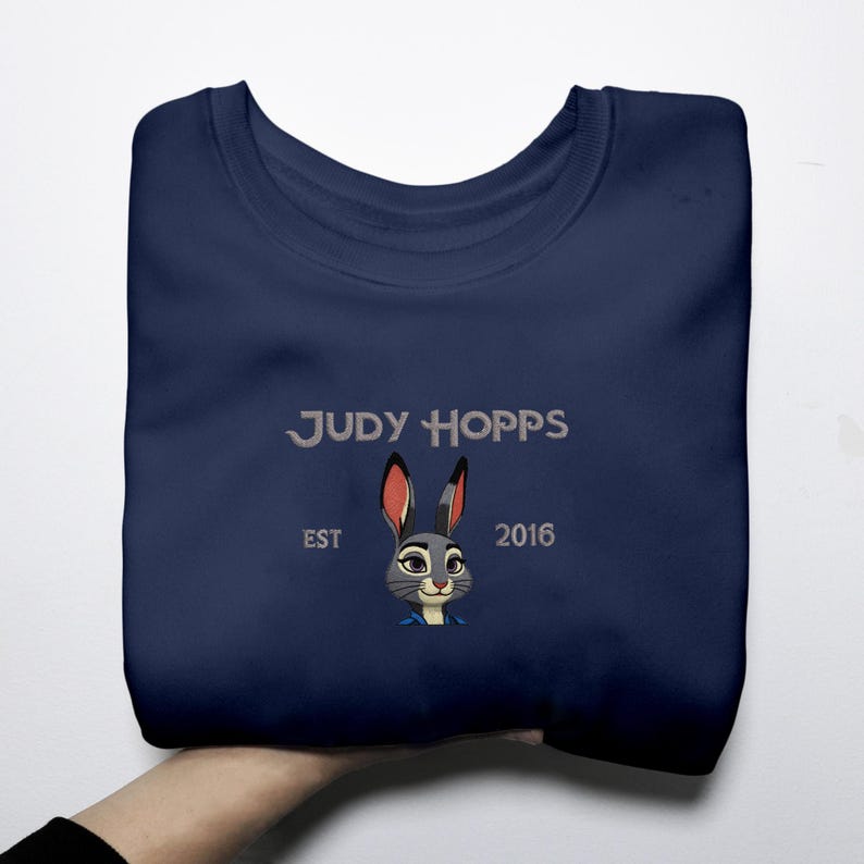 May include: A navy blue sweatshirt with an embroidered design of Judy Hopps, a cartoon rabbit. The text "JUDY HOPPS", "EST", and "2016" are also embroidered. The design is centered on the front of the folded sweatshirt.