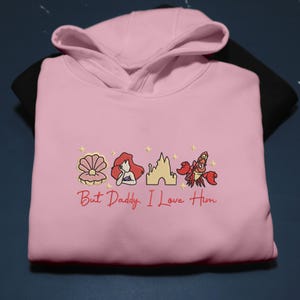 Pode incluir: Um moletom rosa claro com imagens bordadas de uma concha, uma sereia, um castelo e um caranguejo. O texto "But Daddy, I Love Him" &eacute; bordado em vermelho. O moletom est&aacute; em uma superf&iacute;cie preta.