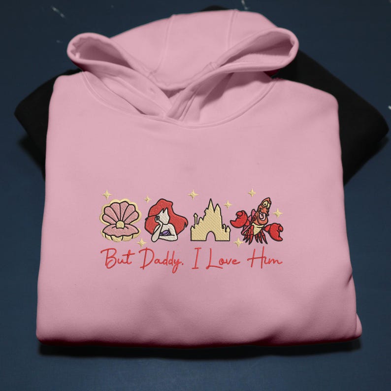 Pode incluir: Um moletom rosa claro com imagens bordadas de uma concha, uma sereia, um castelo e um caranguejo. O texto "But Daddy, I Love Him" &eacute; bordado em vermelho. O moletom est&aacute; em uma superf&iacute;cie preta.