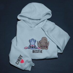 K&ouml;nnte beinhalten: Hellblauer Kapuzenpullover mit Kapuze und langen &Auml;rmeln. Auf der Vorderseite befindet sich ein gesticktes Design mit zwei Zeichentrickfiguren und dem Wort "BESTIE". Der linke &Auml;rmel tr&auml;gt den Namen "CINDY" und ein rotes Herz.