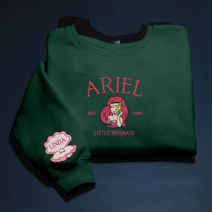 May include: A dark green sweatshirt with the name "ARIEL" in pink script and a cartoon image of Ariel from The Little Mermaid. The sweatshirt also features "EST. 1989" and "LITTLE MERMAID". A pink seashell patch with the name "LINDA" is on the sleeve.