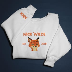 May include: White sweatshirt with an embroidered image of Nick Wilde, a fox character. The sweatshirt has the text "NICK WILDE" above the fox image, with "EST 2016" below. The word "NICK" is also embroidered on the sleeve.