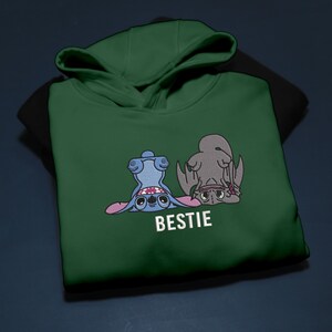 K&ouml;nnte beinhalten: Ein dunkelgr&uuml;ner Kapuzenpullover mit einer Cartoon-Illustration von zwei Figuren, einer blauen und einer grauen, mit dem Wort "BESTIE" in Wei&szlig;. Die Figuren sind auf dem Kopf. Der Kapuzenpullover hat eine Kapuze und ein schwarzes Innenfutter.