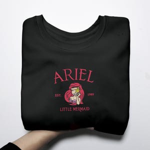 May include: Black sweatshirt with a pink graphic of Ariel from The Little Mermaid. The design includes the name "ARIEL", the text "EST. 1989", and "LITTLE MERMAID". The graphic depicts Ariel with a star.