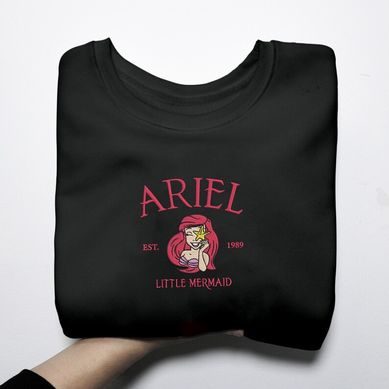May include: Black sweatshirt with a pink graphic of Ariel from The Little Mermaid. The design includes the name "ARIEL", the text "EST. 1989", and "LITTLE MERMAID". The graphic depicts Ariel with a star.