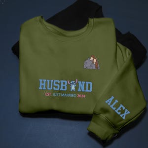 Puede incluir: Sudadera verde oliva con la palabra "HUSBAND" en azul, un personaje de dibujos animados y el texto "EST. JUST MARRIED 2024". Un peque&ntilde;o bordado de una pareja est&aacute; en el pecho. La manga tiene el nombre "ALEX" en azul.