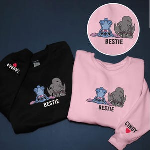 K&ouml;nnte beinhalten: Schwarze und rosa Sweatshirts mit einer Cartoon-Grafik von zwei Figuren, eine blaue und eine graue, mit dem Wort "BESTIE" darunter. Das schwarze Sweatshirt hat "SANDRA" auf dem &Auml;rmel aufgedruckt. Das rosa Sweatshirt hat "CINDY" auf dem &Auml;rmel aufgedruckt.