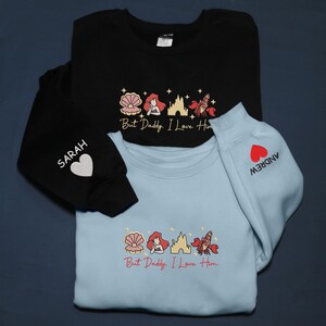 Pode incluir: Duas camisolas, uma preta e outra azul clara, com um desenho bordado de uma concha, uma sereia, um castelo e um caranguejo. O texto "But Daddy, I Love Him" est&aacute; em baixo. Uma manga diz "SARAH" e a outra "ANDREW".