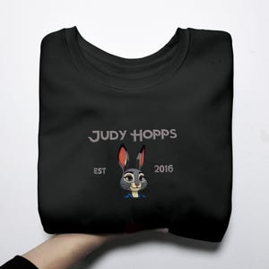 May include: Black crew neck sweatshirt with the text "JUDY HOPPS" and an illustration of Judy Hopps from Zootopia. The design includes "EST 2016". The sweatshirt is a casual style.