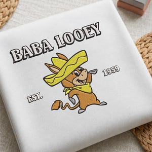Puede incluir: Una sudadera blanca con un bordado de un burro de dibujos animados marrón que lleva un sombrero y un pañuelo amarillos. El texto BABA LOOEY está bordado en negro sobre el personaje, con EST. y 1959 a ambos lados del burro.
