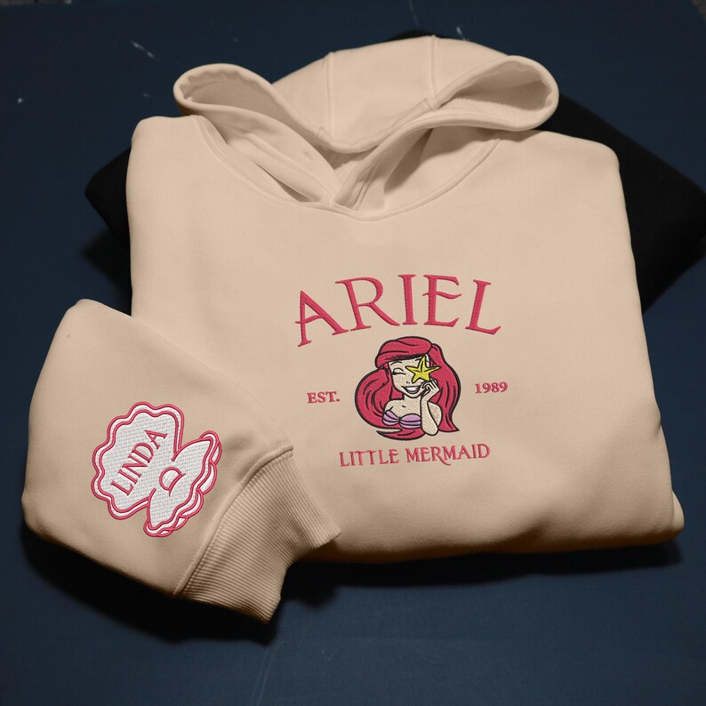 May include: A cream-coloured hoodie with a hood and a matching face mask. The hoodie features the name "ARIEL" in pink script, a Little Mermaid graphic, and the text "EST. 1989 LITTLE MERMAID". The mask has the name "LINDA" embroidered on it.