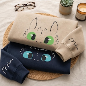 May include: A beige sweatshirt featuring an embroidered green-eyed dragon face and a navy blue sweatshirt with a light blue-eyed dragon face. Both sleeves are embroidered with the name Mike in cursive script, showcasing detailed character designs.
