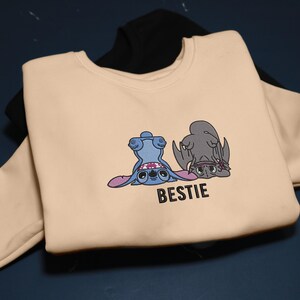 K&ouml;nnte beinhalten: Beiges Sweatshirt mit einem Cartoon-Motiv von zwei Figuren, eine blaue und eine graue, mit dem Wort "BESTIE" darunter. Ein schwarzes Sweatshirt ist teilweise im Hintergrund zu sehen.