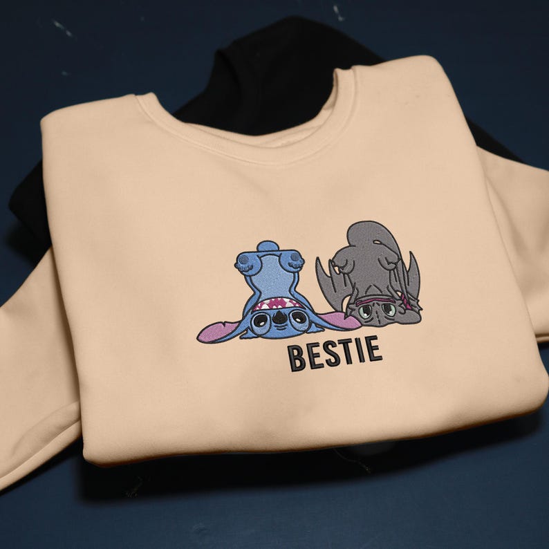 K&ouml;nnte beinhalten: Beiges Sweatshirt mit einem Cartoon-Motiv von zwei Figuren, eine blaue und eine graue, mit dem Wort "BESTIE" darunter. Ein schwarzes Sweatshirt ist teilweise im Hintergrund zu sehen.