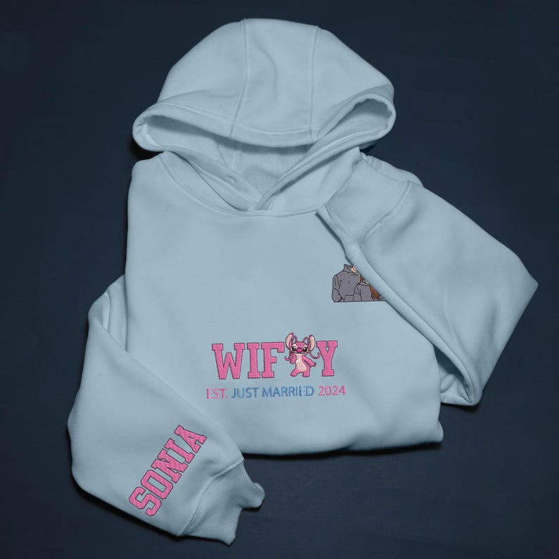 Puede incluir: Una sudadera con capucha azul claro con la palabra "WIFEY" en rosa, un personaje de dibujos animados y el texto "1st JUST MARRIED 2024". La manga tiene el nombre "SONIA" en rosa. La sudadera est&aacute; doblada.