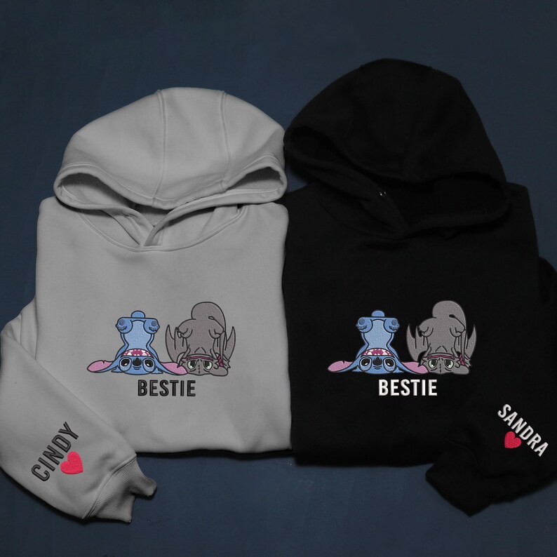 K&ouml;nnte beinhalten: Zwei Kapuzenpullover, einer hellgrau und einer schwarz, mit einem Cartoon-Charakter-Design. Der hellgraue Hoodie hat "Cindy" und ein Herz auf dem &Auml;rmel. Der schwarze Hoodie hat "Sandra" und ein Herz auf dem &Auml;rmel. Beide haben das Wort "Bestie".