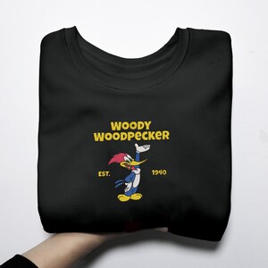 May include: Black sweatshirt featuring a Woody Woodpecker graphic. The design includes the text "WOODY WOODPECKER" in yellow, with an image of the cartoon character. The sweatshirt also has the text "EST. 1940".