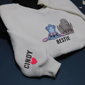 K&ouml;nnte beinhalten: Wei&szlig;es Sweatshirt mit schwarzen &Auml;rmeln, das Cartoon-Figuren und das Wort "BESTIE" zeigt. Auf dem linken &Auml;rmel stehen der Name "CINDY" und ein rotes Herz. Ein l&auml;ssiger Kleidungsstil.