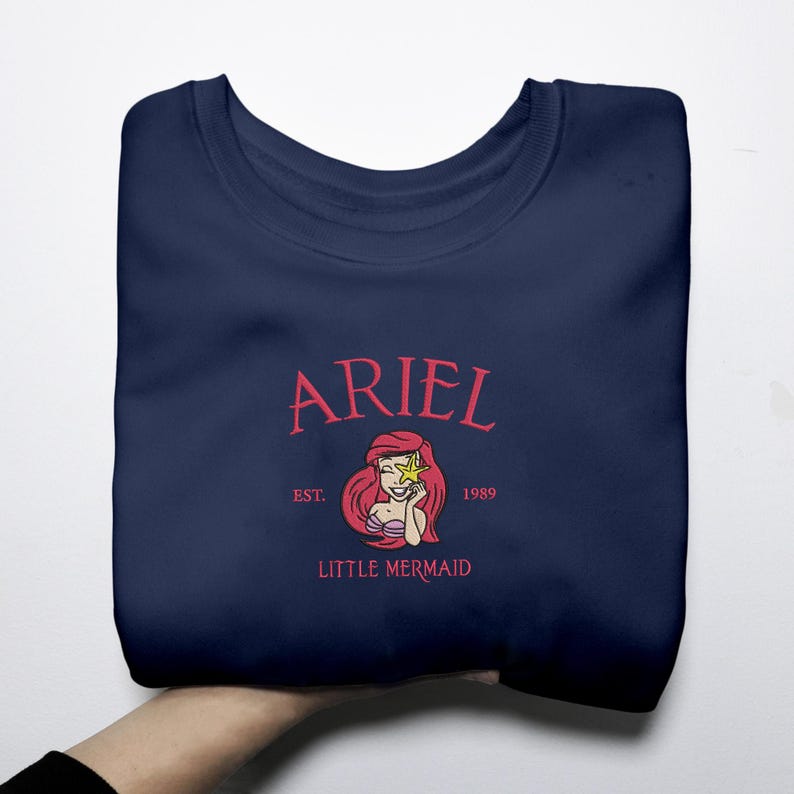 May include: A navy blue sweatshirt with a graphic of Ariel from The Little Mermaid. The design includes the name "ARIEL", the text "EST. 1989", and the words "LITTLE MERMAID". The graphic shows Ariel with red hair and a yellow star.