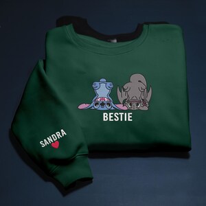 K&ouml;nnte beinhalten: Dunkelgr&uuml;nes Sweatshirt mit einem Cartoon-Motiv von zwei Figuren, eine blaue und eine graue, mit dem Wort "BESTIE" in Wei&szlig;. Der &Auml;rmel zeigt den Namen "SANDRA" und ein rotes Herz.