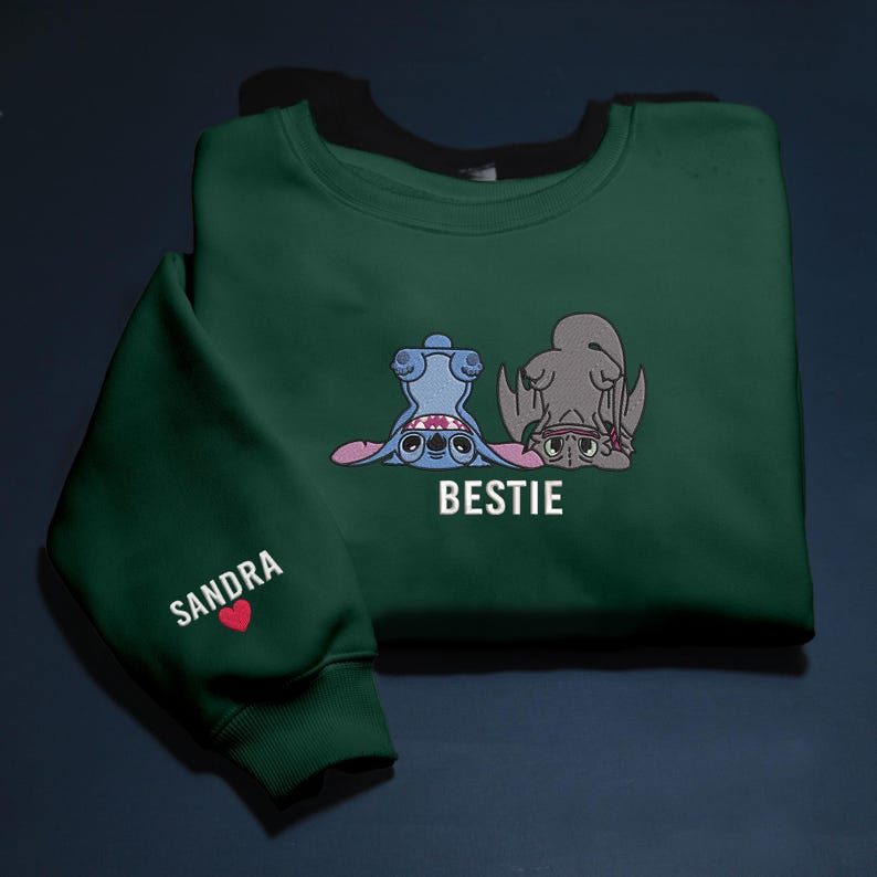 K&ouml;nnte beinhalten: Dunkelgr&uuml;nes Sweatshirt mit einem Cartoon-Motiv von zwei Figuren, eine blaue und eine graue, mit dem Wort "BESTIE" in Wei&szlig;. Der &Auml;rmel zeigt den Namen "SANDRA" und ein rotes Herz.