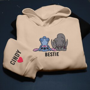 K&ouml;nnte beinhalten: Cremefarbener Kapuzenpullover mit einer blauen Cartoonfigur und einer grauen Cartoonfigur, beide kopf&uuml;ber. Das Wort "BESTIE" steht unter den Figuren. Der linke &Auml;rmel tr&auml;gt den Namen "CINDY" und ein rotes Herz.