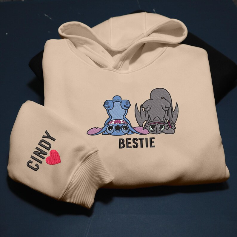 K&ouml;nnte beinhalten: Cremefarbener Kapuzenpullover mit einer blauen Cartoonfigur und einer grauen Cartoonfigur, beide kopf&uuml;ber. Das Wort "BESTIE" steht unter den Figuren. Der linke &Auml;rmel tr&auml;gt den Namen "CINDY" und ein rotes Herz.