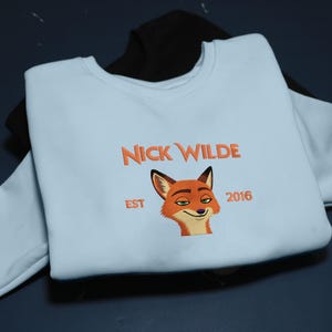 May include: A pale blue sweatshirt with the orange text "NICK WILDE" and a fox illustration. The word "EST" and the number "2016" are also in orange. A black sweatshirt is visible underneath.