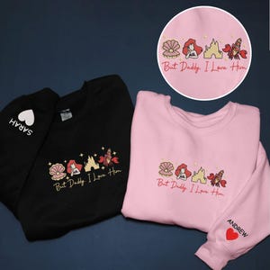 Pode incluir: Camisolas pretas e rosa com desenhos bordados. Os desenhos incluem uma concha, uma sereia, um castelo e um caranguejo, com o texto "But Daddy, I Love Him". Uma camisola tem "SARAH" e um cora&ccedil;&atilde;o, a outra "ANDREW" e um cora&ccedil;&atilde;o.