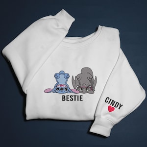 K&ouml;nnte beinhalten: Wei&szlig;es Sweatshirt mit einem Cartoon-Design von zwei Charakteren, einem blauen und einem grauen, mit dem Wort "BESTIE" darunter. Der rechte &Auml;rmel hat den Namen "CINDY" und ein rotes Herz.
