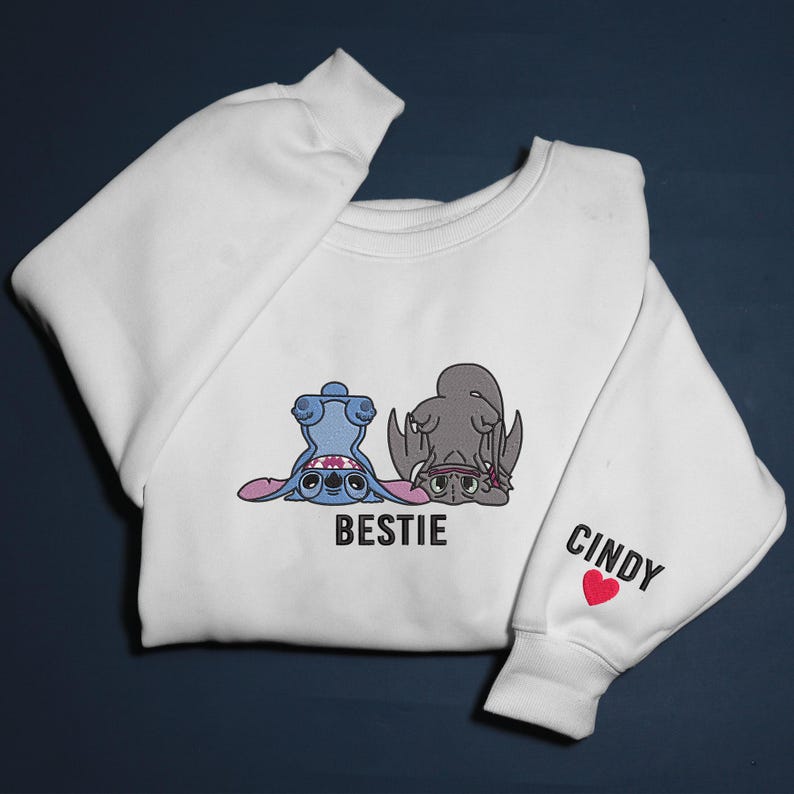 K&ouml;nnte beinhalten: Wei&szlig;es Sweatshirt mit einem Cartoon-Design von zwei Charakteren, einem blauen und einem grauen, mit dem Wort "BESTIE" darunter. Der rechte &Auml;rmel hat den Namen "CINDY" und ein rotes Herz.