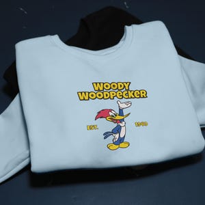 May include: A light blue sweatshirt featuring the Woody Woodpecker cartoon character. The design includes the text "WOODY WOODPECKER" in yellow, the character illustration, and the text "EST. 1940". A black sweatshirt is partially visible underneath.