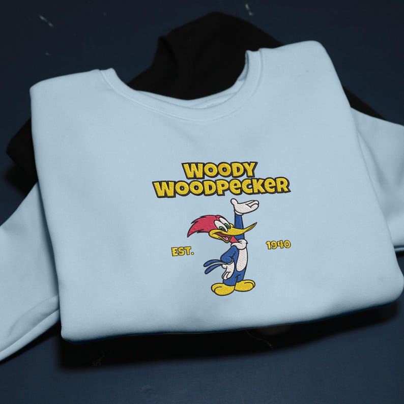 May include: A light blue sweatshirt featuring the Woody Woodpecker cartoon character. The design includes the text "WOODY WOODPECKER" in yellow, the character illustration, and the text "EST. 1940". A black sweatshirt is partially visible underneath.