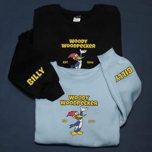 May include: Two sweatshirts, one black and one light blue, featuring the Woody Woodpecker cartoon character. The black sweatshirt has "BILLY" on the sleeve. Both sweatshirts have "WOODY WOODPECKER" and "EST. 1940" printed on them.