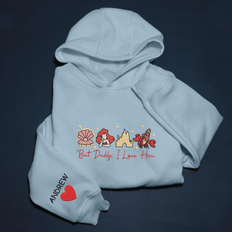 Pode incluir: Um moletom azul claro com capuz e mangas compridas. A frente apresenta um desenho com uma concha, uma mulher ruiva, um castelo e um caranguejo. O texto "But Daddy, I Love Him" est&aacute; abaixo. A manga esquerda tem o nome "ANDREW" e um cora&ccedil;&atilde;o vermelho.