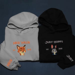 May include: Two hooded sweatshirts, one light grey and one black. The light grey sweatshirt features the text "NICK WILDE" and a fox graphic. The black sweatshirt has "JUDY HOPPS" and a rabbit graphic. Both have "EST 2016".
