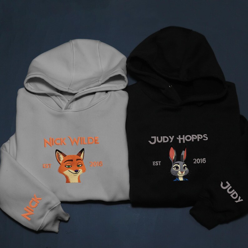 May include: Two hooded sweatshirts, one light grey and one black. The light grey sweatshirt features the text "NICK WILDE" and a fox graphic. The black sweatshirt has "JUDY HOPPS" and a rabbit graphic. Both have "EST 2016".