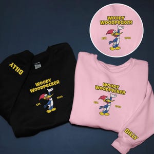 May include: Black and pink sweatshirts featuring the cartoon character Woody Woodpecker. The sweatshirts have the text "WOODY WOODPECKER" and "EST. 1940" printed on the front. The black sweatshirt has "BILLY" printed on the sleeve.