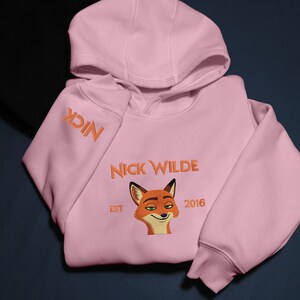 May include: A pale pink hoodie with the name "Nick Wilde" embroidered in orange. The hoodie features an orange fox face graphic and the text "EST 2016". The word "NICK" is embroidered on the left sleeve.