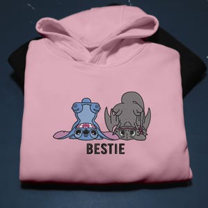 K&ouml;nnte beinhalten: Ein hellrosa Kapuzenpullover mit einem gestickten Design von zwei Zeichentrickfiguren, eine blaue und eine graue, in einer spielerischen Pose. Das Wort "BESTIE" ist darunter gedruckt. Der Kapuzenpullover liegt auf einem schwarzen Kleidungsst&uuml;ck.