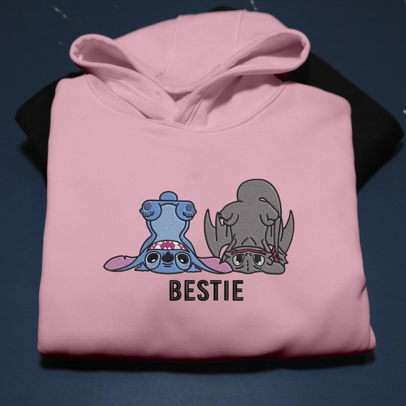 K&ouml;nnte beinhalten: Ein hellrosa Kapuzenpullover mit einem gestickten Design von zwei Zeichentrickfiguren, eine blaue und eine graue, in einer spielerischen Pose. Das Wort "BESTIE" ist darunter gedruckt. Der Kapuzenpullover liegt auf einem schwarzen Kleidungsst&uuml;ck.