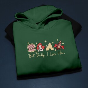 Pode incluir: Um moletom com capuz verde escuro com imagens bordadas de uma concha, uma sereia, um castelo e um caranguejo. O texto "But Daddy, I Love Him" &eacute; bordado em dourado abaixo.